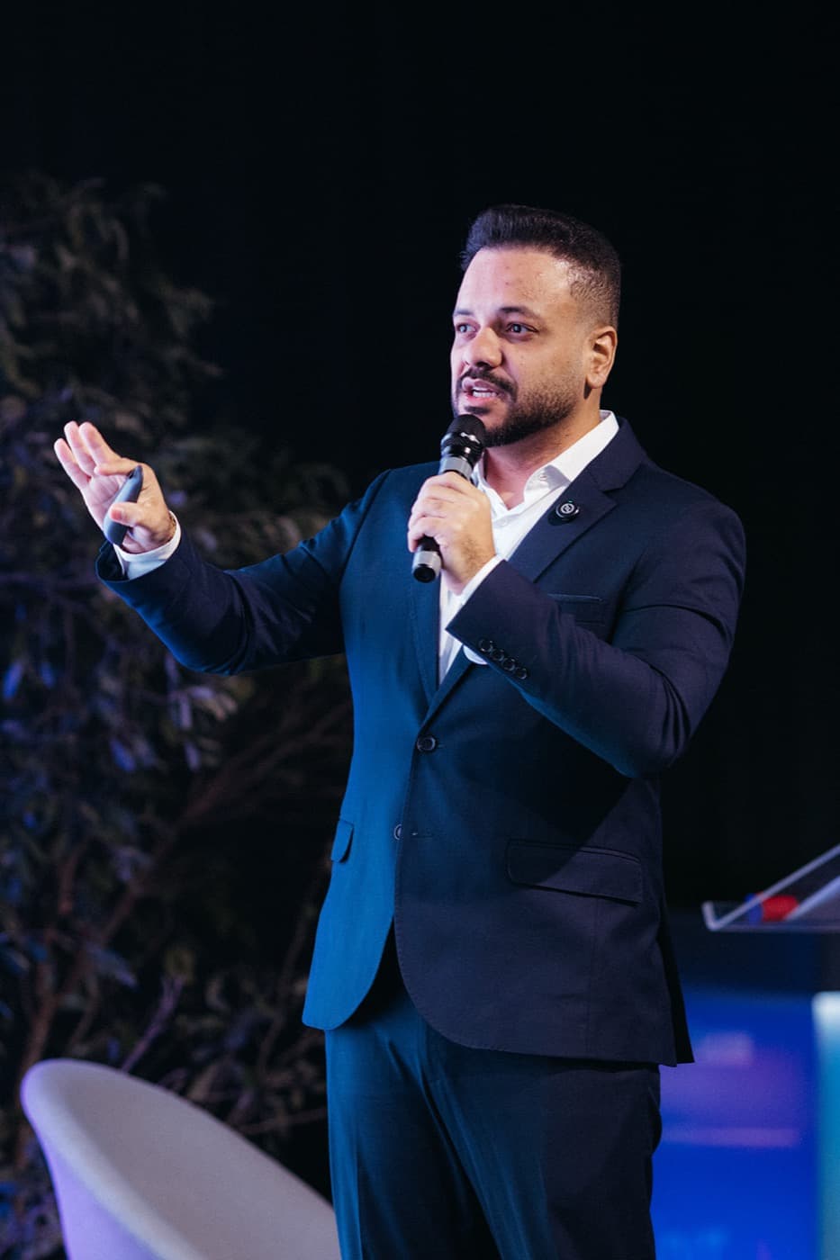 Anderlan Oliveira palestrando em evento, de terno azul, com microfone